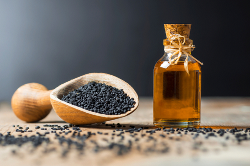 Wooden spoon of black cumin seeds sits next to corked bottle of black seed oil